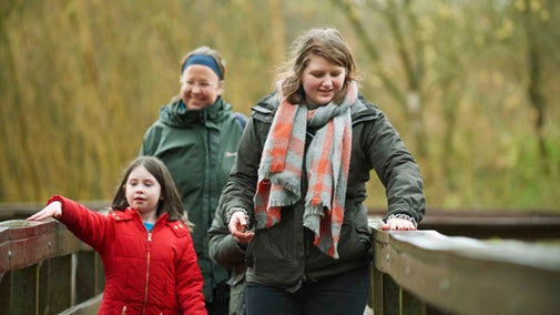 A family are crossing a sturdy wooden bridge at Winkworth Arboretum, Surrey, and there are trees in soft focus in the background.